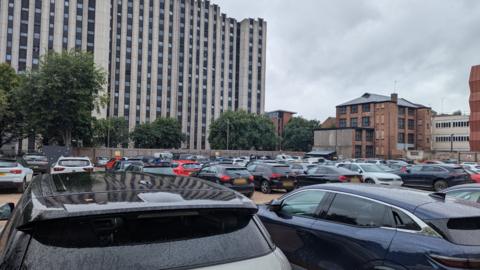 Rows of parked cars in front of a large grey building behind the car park
