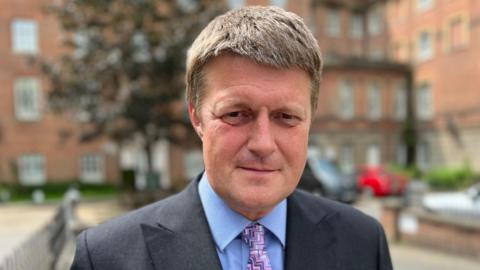 Head and shoulders image of Richard Clewer in a dark suit with a blue shirt and pink, zig, zag pattern tie