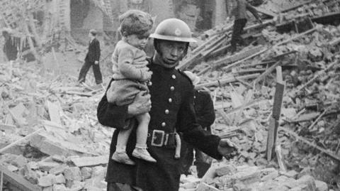 Black and white photo of firefighter wearing uniform and metal helmet carrying a small child across bomb wreckage