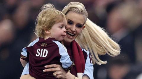 Kelly Osbourne, who has long blonde hair that is waving behind her, is holding a young boy with blonde hair in her arms and smiling. The boy is wearing an Aston Villa shirt that says SID on the back
