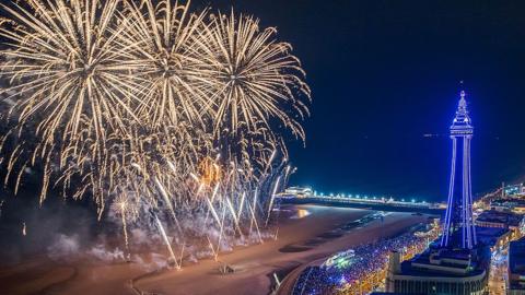 Aerial view of the fireworks display on the beach beside Blackpool's North pier with crowds of people watching on the promenade in the evening with the Blackpool Illuminations on and Blackpool Tower lit up purple in the background to the right.