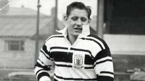 A black and white photo of a man playing rugby league. He is wearing a black and white hooped shirt with a white collar. A badge on the chest depicts three crowns and has the word "Hull" in capital lettering.
