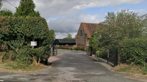 A driveway with two black gates, and hedges either side. Along the drive and to the right a red brick building can be seen, and further beyond that another building that is all clad in black metal