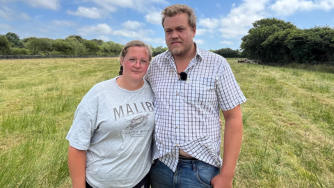 A man and woman stand side by side in an empty grassy field. Trees are visible in the background and there is a blue sky with clouds.
