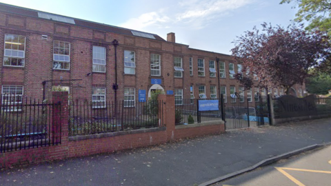 A red brick building with railings outside. There are trees and a blue school sign.