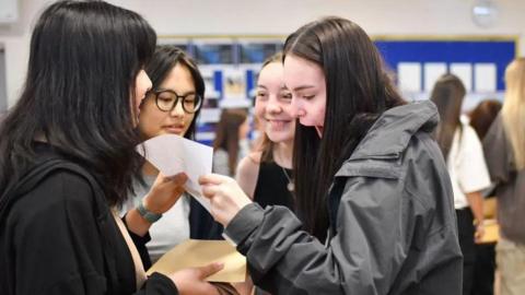 A group of female students stand together looking at results. One is smiling whilst another has her mouth open in shock whilst looking at the paper. 