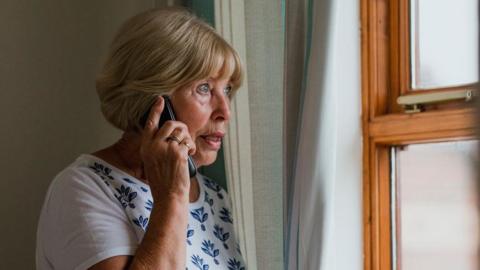 A woman with blond hair and a white patterned top holding a mobile phone to her ear and looking out of a wood-framed window