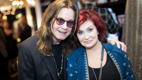 Ozzy Osbourne and Sharon Osbourne. He has long brown hair and is wearing a black jacket and glasses and Sharon is wearing a blue jacket and has redish brown hair.
