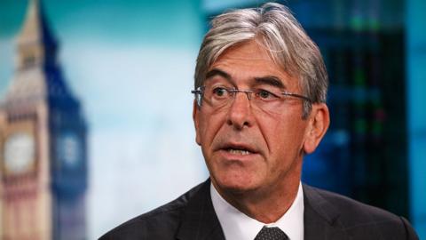 Michael Ward, a man with short dark grey hair wearing wire-frame glasses, looks to his left, against a blueish background with an image of Big Ben out of focus, during a Bloomberg TV interview in 2016.