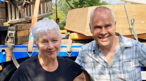 Anna Kaye poses with her arms around her son Jonathan Allen in front of a trailer taking a boat away. They are smiling in the sunshine. She wears a blue t-shirt and Mr Allen wears a light blue checked shirt.