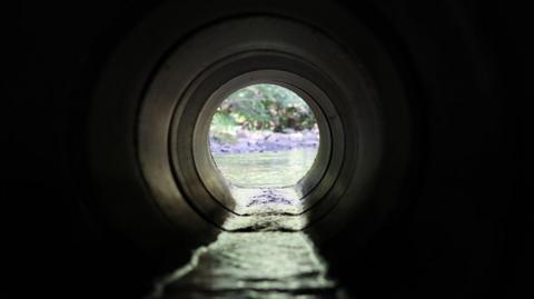 View through a round concrete tunnel carrying a stream beneath a road - stock photo