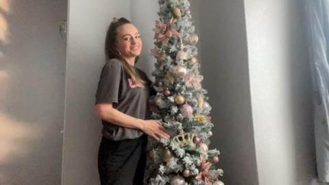 Sally-Anne Long, a woman with her hair tied back wearing a grey t-shirt, stands next to a Christmas tree decorated with gold and red ornaments.