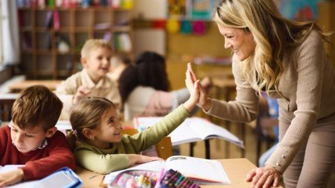 A young teacher is leaning over the desk of two under-10 pupils and is high-fiving the nearest child. Both are smiling.