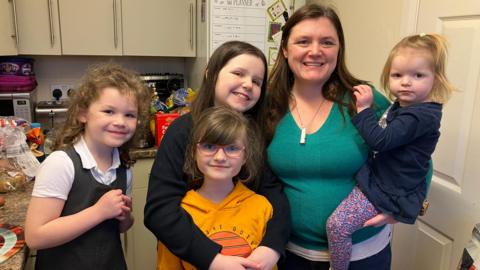 Patricia has long dark hair, a green top and is carrying a small girl on her left hip. Two her right three older girls smile at the camera. They are all standing in a kitchen.
