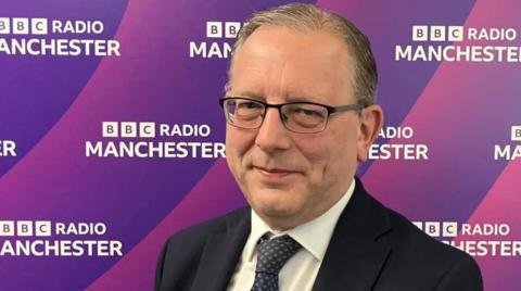 Michael Winstanley, wearing glasses and a black suit, looks at the camera