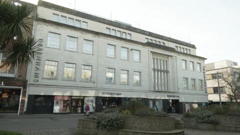 Looking at the Debenhams building's exterior on the High Street in Plymouth. The building is large and cream in colour. The Debenhams logo appears five times on the front of the building. The street is quiet/