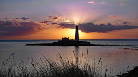 The silhouette of a lighthouse is illuminated by the rising sun. Rays of orange light break through the clouds. In the foreground, grass is swaying in the wind.