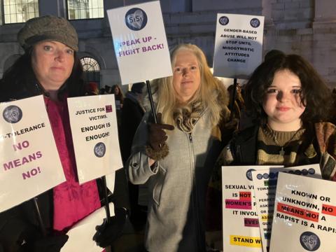 Three women at the protest supporting Nikita Hand - they are standing side by side holding protest signs, including some saying 'no means no'. The women on the left is wearing a cap and pink scarf. The women in the middle is wearing a grey coat and scarf and has shoulder-length blond hair. The women on the right, who is younger, is wearing a striped jumper and has short brown hair. 