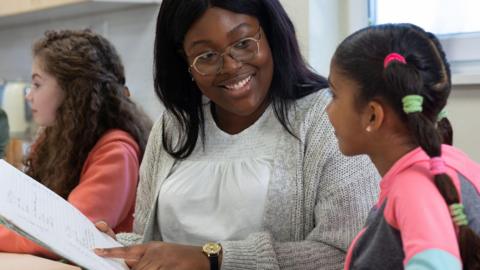 A woman is wearing a grey cardigan and glasses and holding a book while talking to a young girl in pigtails. A third girl can be seen in the background looking away