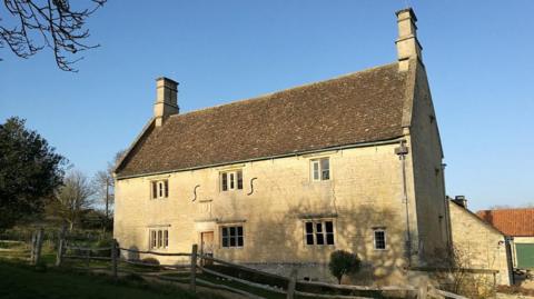 A two floor old house with yellow stone and a brown roof. A fence is located in front of it and greenery and trees can be seen to the left. There are three windows at the top and three at the bottom.