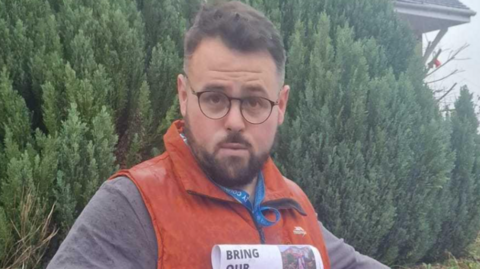A man, wearing glasses, in an orange top with dark hair and a beard is looking at the camera, with bushes behind him.