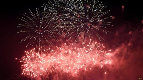 A firework display at night. There are white fireworks at the top, and pink ones below lighting up the school.