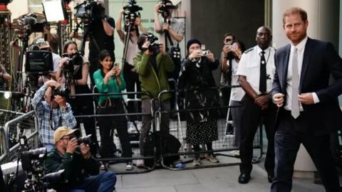 Prince Harry leaves the court during his hacking case against the Daily Mirror. He wears a dark coloured suit, white shirt and tie. His barrister David Sherborne, also dressed in a dark suit is on his left. A crowd of photographers are behind a metal barrier