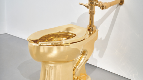 A solid gold toilet fixed on a white wall and a grey floor.