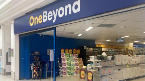 The exterior of One Beyond in The Meadow shopping centre, Chelmsford. Above the wide doorway is a bright blue sign saying One Beyond. Within the shop are stacks of kitchen or loo roll, in brand plastic wrapping, and rows of clear plastic storage boxes. Beyond that is a fridge with rows containing bottles. 