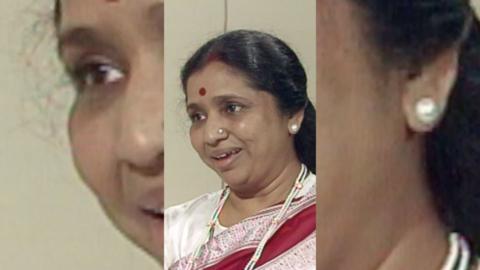 A woman, Asha Bhosle, with dark hair smiling. She is wearing a white, red and pink top.