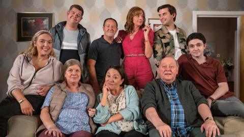 A group of eight people gathered in a living room setting. Four individuals are seated on a couch in the foreground, while four others stand behind them. The background features patterned wallpaper with a leaf design in muted tones.