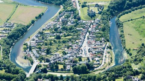 An aerial view of the village of Warkworth in Northumberland. It is surrounded by a river and countryside. There is a castle in the village.