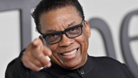 Herbie Hancock pointing straight at the camera. He is wearing black glasses and a collarless black shirt.