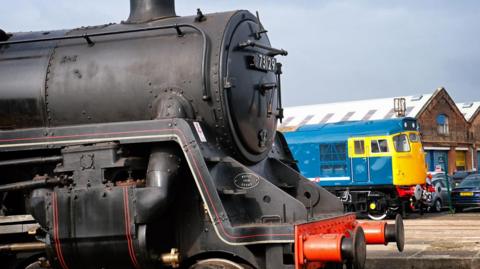A black steam locomotive is parked in the foreground, with a blue and yellow train and brick industrial buildings visible in the background


