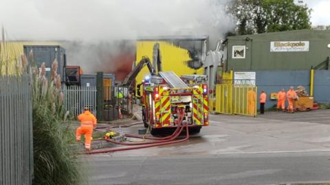 A fire engine is parked at the entrance to an industrial site. A yellow warehouse is on fire with white smoke pouring from the entrance. Part of the cladding has changed colour. Men in orange high viz clothing watch on from both sides of the fire engine. 