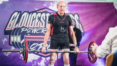 A man in his 80s lifts a barbell on stage, in front of a neon purple backdrop. He is wearing black shorts, a black T-shirt, and a black and red vest. He has receding grey hair. There is a woman standing behind him and someone is sat in the audience clapping.