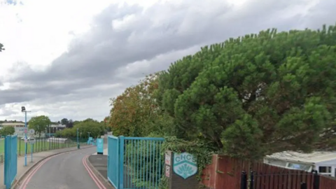 Blue school gates are open with a road leading to the school. The school's logo can be seen to the right of the gates by a wooden fence. There are tree also on the right side with school fields on the left.