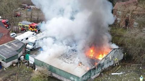 A drone shot of a single-storey brick-built factory unit on fire, surrounded by countryside and two homes