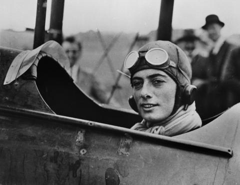 Winifred Spooner, english aviator, sits in a plane preparing to take off. The black and white image shows her sat in the cockpit, with a leather protective helmet on and goggles rested on her forehead. Winnifred is looking slightly off camera.