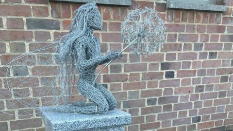 A close-up of a figure on top of a memorial statue. Made of interwoven strands of grey material, it shows a woman with long hair kneeling and holding a dandelion which is about to shed its seeds.