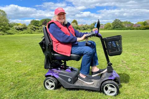 Elderley woman wearing red coat and cap whilst sat on mobility scooter in a field