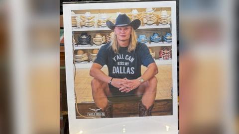 Man in cowboy hat and 'Y'All Can Kiss My Dallas' shirt in front of shelves of cowboy hats