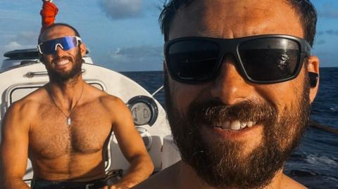 Two men with beards and wearing sunglasses are smiling at the camera. Mr Mason is in the foreground and on the photo a pole, containing a flag, is directly above and behind Mr Mattock's head. The sea is behind the rowing boat they are in.