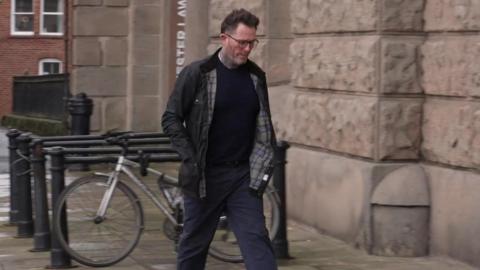A man in blue trousers, a blue jumper and a black Barbour jacket walking towards a court.