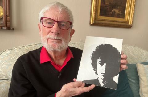 Alan Snook, a man with white hair and a white beard, holds a black and white picture of Bob Dylan. He is sitting on a sofa as he holds the picture