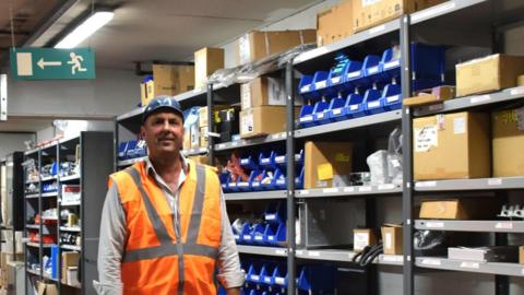 Darren Milligan in a hardhat and high-viz vest at Heaton Train Care Centre in Newcastle. To his left are rows of metal shelves containing boxes and parts.