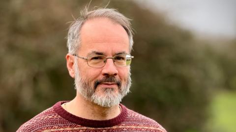 A man stands outside with a large hedge blurred in the background. He is wearing wire rimmed glasses, has short grey hair and a beard. He is wearing a patterned red jumper.
