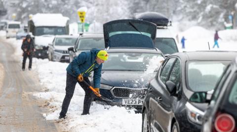 A person in winter clothing shovels snow away from a car on a congested, snow-covered road, with several vehicles stuck in traffic and heavy snowfall visible in a mountainous or rural setting.
