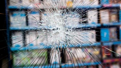 A smashed window of a shop, with the goods seen out of focus behind it. The glass has been smashed into shards but has not fallen out of the window.