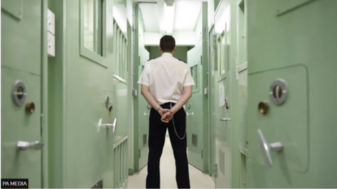 A prison officer, wearing a white shirt and black trousers, stands with his back to the camera. There are pale green prison doors to the left and right of him